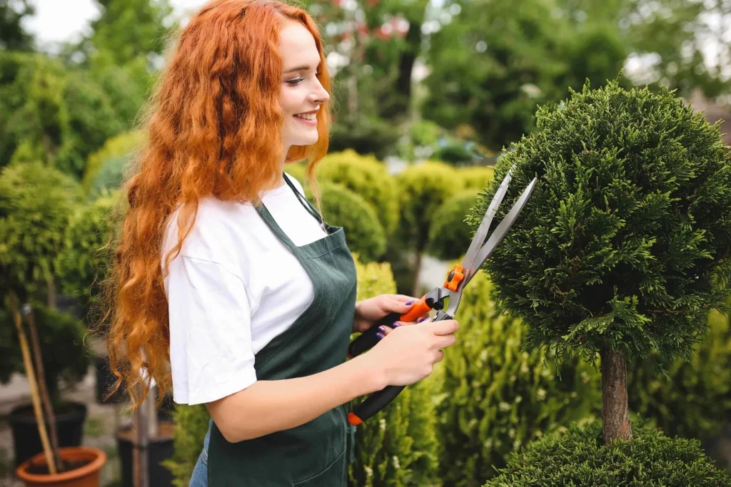Smiling lady with red curly hair standing with apron holding garden scissors while working outdoors